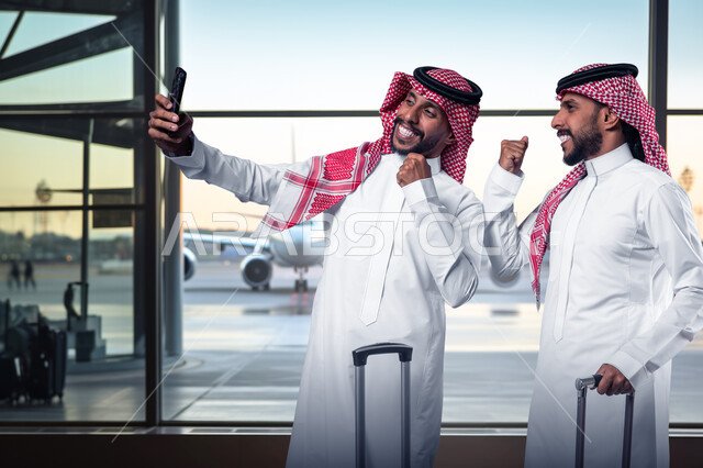 التقاط صور السيلفي التذكارية، مفهوم السياحة والقيام بالجولات الترفيهية في وقت الإجازة، صديقان عربيان خليجيان سعوديان يرتديان الشماغ والثوب التقليدي يمسكان حقائب السفر يقومان باستخدام الهاتف المحمول، الاستعداد لبدء الرحلة في العطلة الصيفيه بإيماءات الحماس والسرور