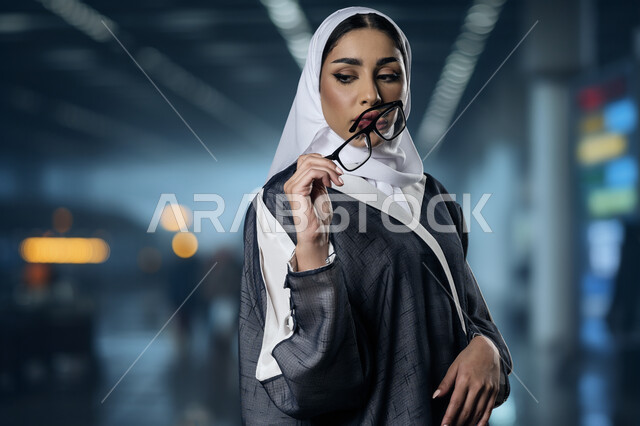 ألبسة نسائية عصرية وأنيقة، صورة مقربة لامرأة عربية خليجية سعودية محجبة ترتدي العباءة تحمل بيدها النظارة الطبية تنظر الى شيء ما بإيماءات الثقة بالنفس، مفهوم الموضة والأناقة والاهتمام بالمظهر الخارجي، تعابير النعومة والأنوثة