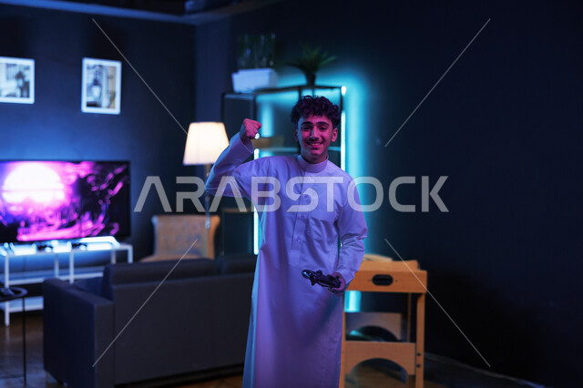 Integration and interaction with video games, a young Saudi Arabian Gulf man wearing traditional clothing standing in his room holding a joystick looking at the camera raising his hand up with gestures of enthusiasm and pleasure, modern entertainment electronic devices and technologies