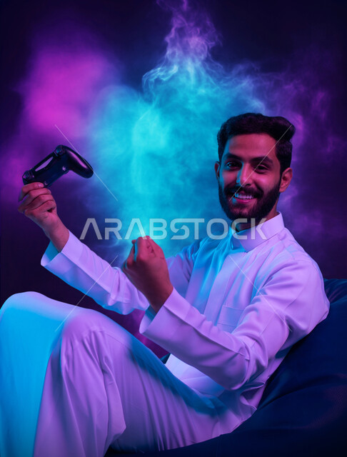 تعابير الاندماج والتفاعل، بورتريه لشاب عربي سعودي خليجي يرتدي الثوب التقليدي يجلس على كرسي مريح يحمل بيده عصا التحكم يقوم باللعب في ألعاب الفيديو ينظر الى الكاميرا بإيماءات الحماس والسرور، اجهزة وتقنيات الكترونية حديثة ترفيهيه, خلفية ملونة