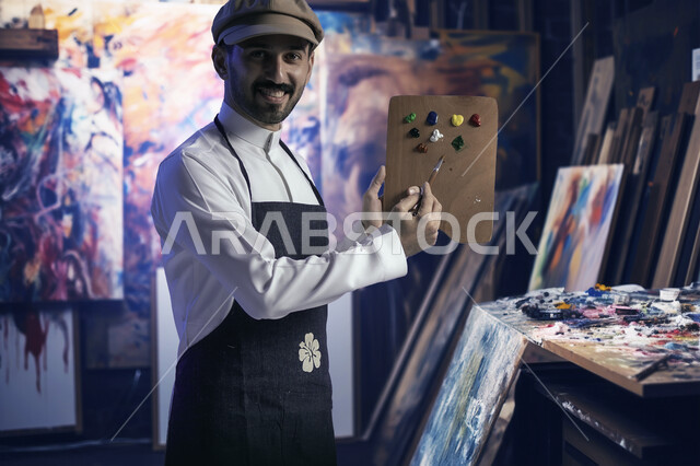 الشغف في ممارسة الفنون اليدوية و الهوايات المفضلة, صورة مقربة لرسام عربي خليجي سعودي يرتدي الثوب التقليدي و مريلة الرسم يحمل بيده لوح الالوان ينظر الى الكاميرا بإيماءات السرور, مفهوم الموهبة و الإبداع, أنشطة ترفيهية فنية ممتعة