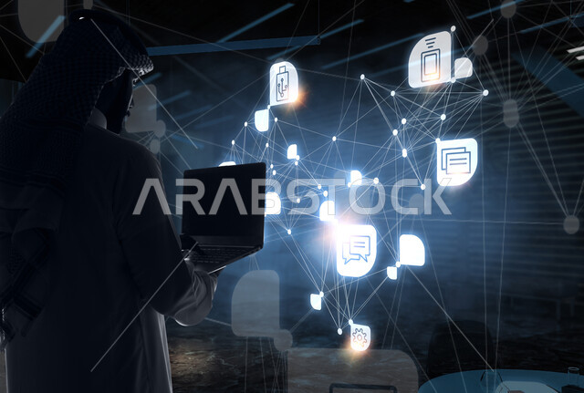 مفهوم التحول الرقمي، دمج التكنولوجيا المتطورة مع العمل، استخدام التقنيات الحديثة، صورة من الخلف لرجل أعمال عربي خليجي سعودي يرتدي الشماغ والثوب التقليدي يمسك بيده جهاز اللابتوب المحمول يقف امام شاشة إلكترونية بأيقونات هولوجرامية مضيئة، رؤية مستقبلية تكنولوجية