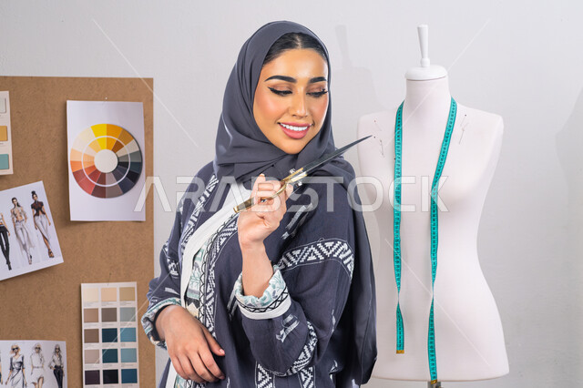 استعمال أدوات ومعدات الخياطة، اختيار أجود أنواع الأقمشة لصناعة الأثواب، احتراف و إتقان صنع الملابس السعودية، حرف ومهن يدوية، بورتريه مقربة لخياطة عربية خليجية سعودية محجبة ترتدي العباءة تحمل بيدها المقص، تعابير السرور والثقة بالنفس، خلفية رمادية