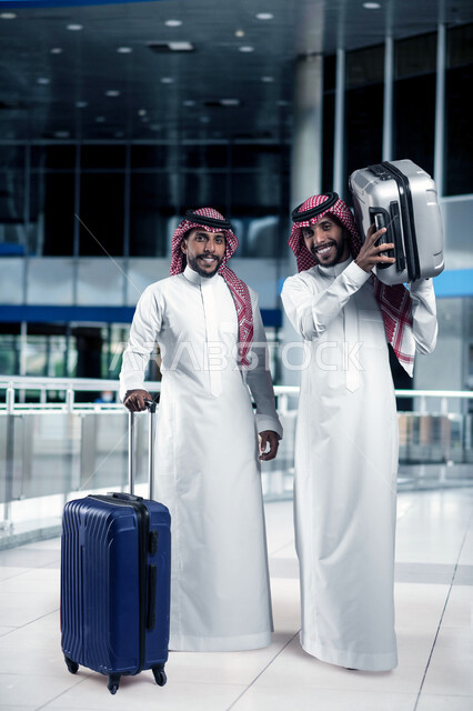 The concept of tourism and leisure tours during vacation time, two Saudi Arabian Gulf friends wearing traditional shemagh and thobe carrying suitcases preparing to go on a trip, looking at the camera with gestures of enthusiasm and pleasure, full body image