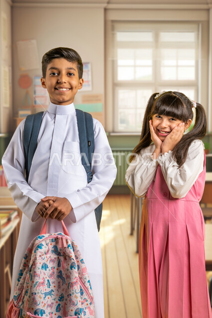 Back to school, two Saudi Arabian Gulf students wearing school uniforms carrying school bags standing inside the classroom looking at the camera with gestures of joy and happiness, preparing for the new school year, a girl putting her hand on her face with expressions of enthusiasm and activity