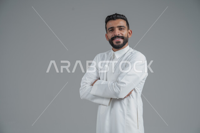 الاهتمام بالمظهر الخارجي والأناقة، مفهوم الرجولة والثقة بالنفس، بورتريه لرجل عربي خليجي سعودي يرتدي الثوب التقليدي ينظر الى الكاميرا بتعابير السرور، الوقوف باستقامة وثبات بايدي متقاطعة،  خلفية رمادية