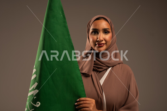 ترسيخ جذور الانتماء الوطني، بورتريه مقرب لامرأة عربية خليجية سعودية محجبة ترتدي عباءة بنية تحمل علم المملكة بإيماءات السرور والسعادة، الاحتفال بيوم العلم 11 مارس، احياء ذكرى العيد الوطني السعودي 23 سبتمبر، خلفية رمادية