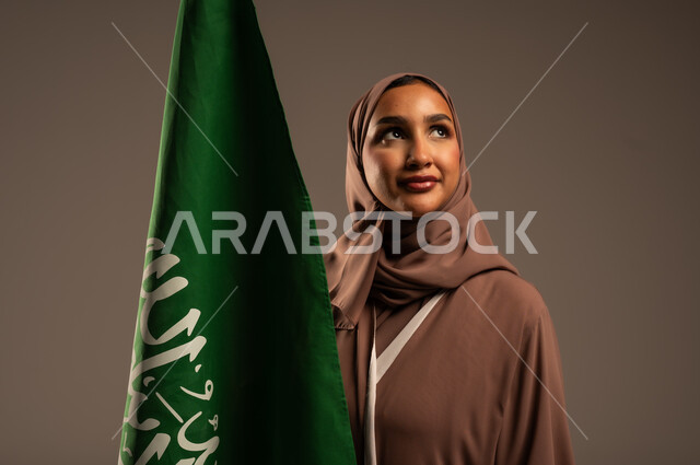 ترسيخ جذور الانتماء الوطني، بورتريه مقرب لامرأة عربية خليجية سعودية محجبة ترتدي عباءة بنية تحمل علم المملكة بإيماءات السرور والسعادة، الاحتفال بيوم العلم 11 مارس، احياء ذكرى العيد الوطني السعودي 23 سبتمبر، خلفية رمادية