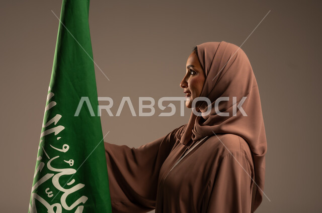 ترسيخ جذور الانتماء الوطني، بورتريه مقرب لامرأة عربية خليجية سعودية محجبة ترتدي عباءة بنية تحمل علم المملكة بإيماءات السرور والسعادة، الاحتفال بيوم العلم 11 مارس، احياء ذكرى العيد الوطني السعودي 23 سبتمبر، خلفية رمادية