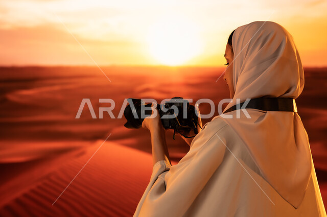 ممارسة الهواية المفضلة بشغف، مصورة محترفة تقوم بالتقاط الصور التذكارية باستخدام التقنيات الحديثة، الرمال الذهبية الناعمة, صورة من الخلف لشابة عربية خليجية سعودية ترتدي عباءة ملونة تقف في المناطق الصحراوية تحمل بيديها كاميرا عالية الدقة، لقطات فنية فوتوغرافية