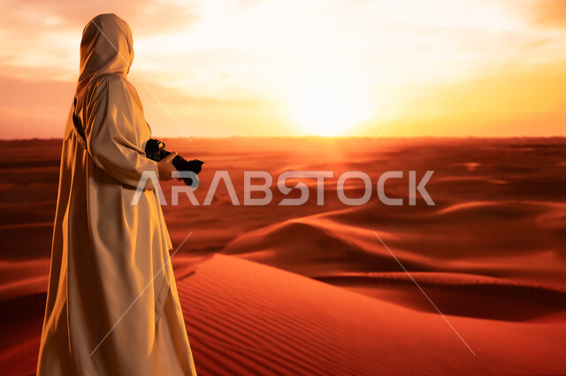 ممارسة الهواية المفضلة بشغف، مصورة محترفة تقوم بالتقاط الصور التذكارية باستخدام التقنيات الحديثة، الرمال الذهبية الناعمة, صورة من الخلف لشابة عربية خليجية سعودية ترتدي عباءة ملونة تقف في المناطق الصحراوية تحمل بيديها كاميرا عالية الدقة، لقطات فنية فوتوغرافية