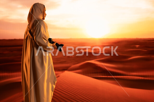 Passionately practicing her favorite hobby, a professional photographer taking souvenir photos using modern techniques, soft golden sand, a photo from the back of a young Saudi Arabian Gulf woman wearing a colorful abaya standing in the desert areas holding a high-resolution camera in her hands, artistic photographic shots