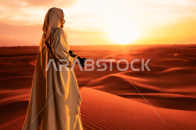 ممارسة الهواية المفضلة بشغف، مصورة محترفة تقوم بالتقاط الصور التذكارية باستخدام التقنيات الحديثة، الرمال الذهبية الناعمة, صورة من الخلف لشابة عربية خليجية سعودية ترتدي عباءة ملونة تقف في المناطق الصحراوية تحمل بيديها كاميرا عالية الدقة، لقطات فنية فوتوغرافية