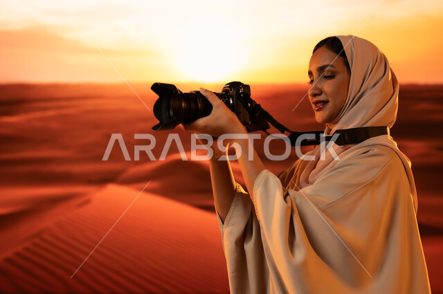 لقطات فنية فوتوغرافية، ممارسة الهواية المفضلة بشغف، مصورة محترفة تقوم بالتقاط الصور التذكارية باستخدام التقنيات الحديثة، الرمال الذهبية الناعمة, صورة من الجانب لشابة عربية خليجية سعودية ترتدي عباءة ملونة تقف في المناطق الصحراوية تحمل بيديها كاميرا عالية الدقة