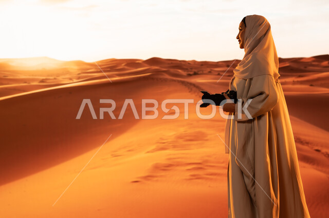 Artistic photography shots, practicing a favorite hobby with passion, a professional photographer taking souvenir photos using modern techniques, soft golden sand, a side view of a young Saudi Arabian Gulf woman wearing a colorful abaya standing in the desert areas holding a high-resolution camera in her hands
