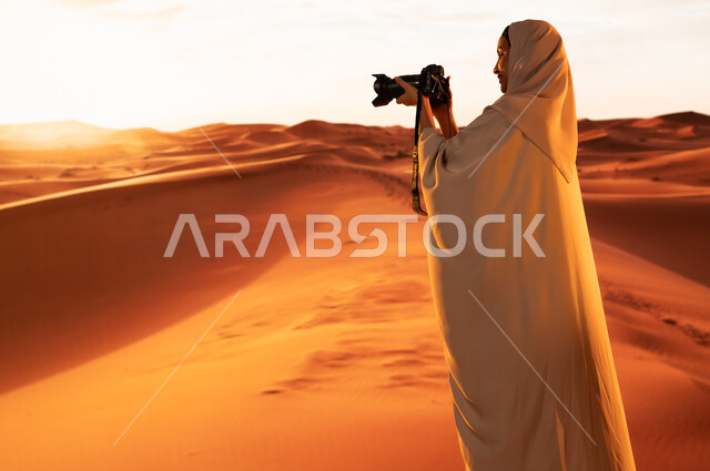Artistic photography shots, practicing a favorite hobby with passion, a professional photographer taking souvenir photos using modern techniques, soft golden sand, a side view of a young Saudi Arabian Gulf woman wearing a colorful abaya standing in the desert areas holding a high-resolution camera in her hands