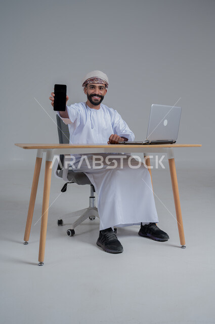 مفهوم ادارة الاعمال عن بعد، مهن ووظائف مكتبيه عمانية، بورتريه لشاب عربي خليجي عماني يرتدي الدشداشة والعمامة يجلس خلف طاولة خشبية وامامه حاسوب يقوم بعرض شاشة سوداء فارغة، استخدام الأجهزة الحديثة والتقنيات المتطورة، خلفية بيضاء