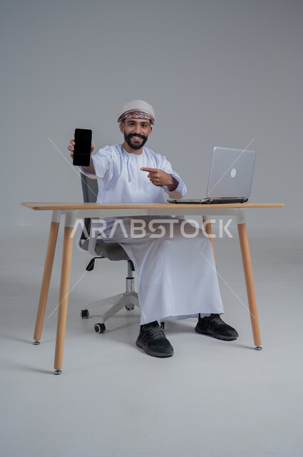 مفهوم ادارة الاعمال عن بعد، مهن ووظائف مكتبيه عمانية، بورتريه لشاب عربي خليجي عماني يرتدي الدشداشة والعمامة يجلس خلف طاولة خشبية وامامه حاسوب يقوم بعرض شاشة سوداء فارغة، استخدام الأجهزة الحديثة والتقنيات المتطورة، خلفية بيضاء