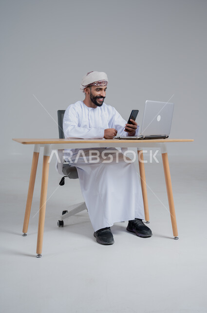 مفهوم ادارة الاعمال عن بعد، مهن ووظائف مكتبيه عمانية، بورتريه لشاب عربي خليجي عماني يرتدي الدشداشة والعمامة يجلس خلف طاولة خشبية وامامه حاسوب يحمل بيده هاتف محمول، استخدام الأجهزة الحديثة والتقنيات المتطورة، خلفية بيضاء