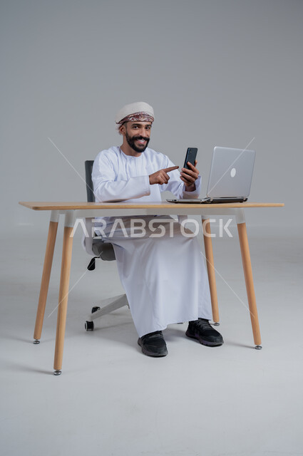 مفهوم ادارة الاعمال عن بعد، مهن ووظائف مكتبيه عمانية، بورتريه لشاب عربي خليجي عماني يرتدي الدشداشة والعمامة يجلس خلف طاولة خشبية وامامه حاسوب يحمل بيده هاتف محمول، استخدام الأجهزة الحديثة والتقنيات المتطورة، خلفية بيضاء