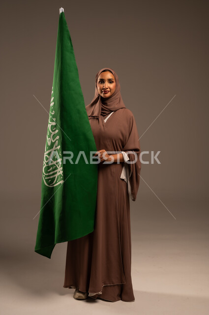 الاحتفال بيوم العلم 11 مارس، ترسيخ جذور الانتماء الوطني، بورتريه لامرأة عربية خليجية سعودية محجبة ترتدي عباءة بنية تحمل علم المملكة بإيماءات السرور والسعادة، احياء ذكرى العيد الوطني السعودي 23 سبتمبر، الطول الكامل للجسم، خلفية رمادية