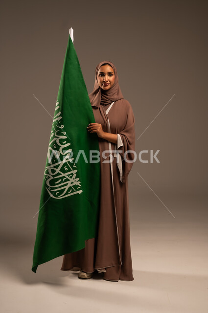 الاحتفال بيوم العلم 11 مارس، ترسيخ جذور الانتماء الوطني، بورتريه لامرأة عربية خليجية سعودية محجبة ترتدي عباءة بنية تحمل علم المملكة بإيماءات السرور والسعادة، احياء ذكرى العيد الوطني السعودي 23 سبتمبر، الطول الكامل للجسم، خلفية رمادية