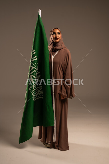 الاحتفال بيوم العلم 11 مارس، ترسيخ جذور الانتماء الوطني، بورتريه لامرأة عربية خليجية سعودية محجبة ترتدي عباءة بنية تحمل علم المملكة بإيماءات السرور والسعادة، احياء ذكرى العيد الوطني السعودي 23 سبتمبر، الطول الكامل للجسم، خلفية رمادية