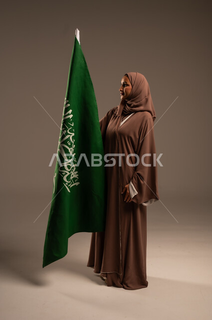 Celebrating Flag Day March 11, consolidating the roots of national belonging, portrait of a veiled Saudi Arabian Gulf woman wearing a brown abaya holding the Kingdom's flag with gestures of joy and happiness, commemorating Saudi National Day September 23, full body length, gray background