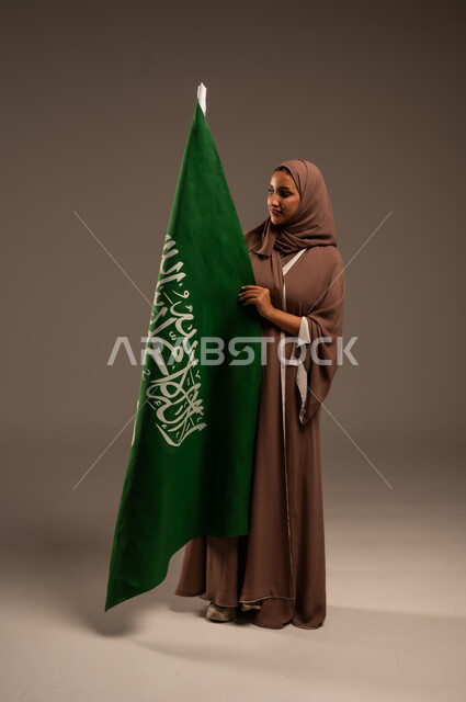 احياء ذكرى العيد الوطني السعودي 23 سبتمبر، الاحتفال بيوم العلم 11 مارس، ترسيخ جذور الانتماء الوطني، بورتريه لامرأة عربية خليجية سعودية محجبة ترتدي عباءة بنية تحمل علم المملكة بإيماءات السرور والسعادة، الطول الكامل للجسم، خلفية رمادية