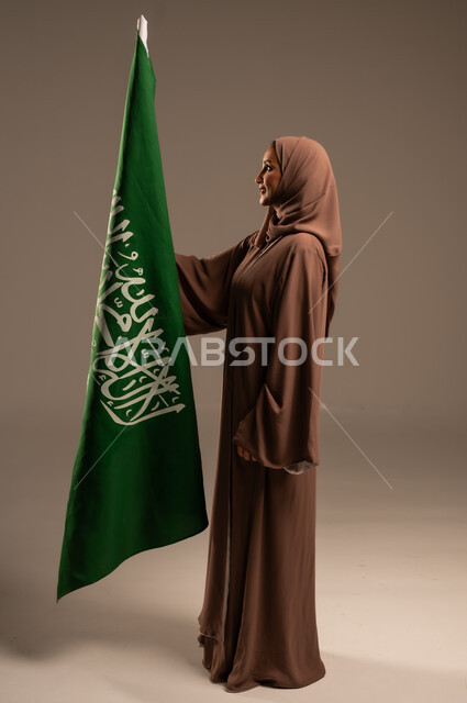 Commemorating Saudi National Day September 23, Celebrating Flag Day March 11, Establishing the Roots of National Belonging, Portrait of a Veiled Saudi Arabian Gulf Woman Wearing a Brown Abaya Holding the Kingdom's Flag with Gestures of Joy and Happiness, Full Body Length, Grey Background