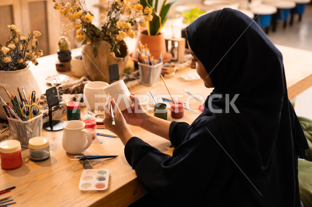 العمل في صناعة الخزفيات, تأسيس الدولة السعودية 1727 م, ذكرى اليوم الوطني السعودي23 سبتمبر, فن الرسم والتلوين على الخزف، امرأة عربية خليجية سعودية محجبة ترتدي العباءة السوداء تقوم بالرسم على السيراميك