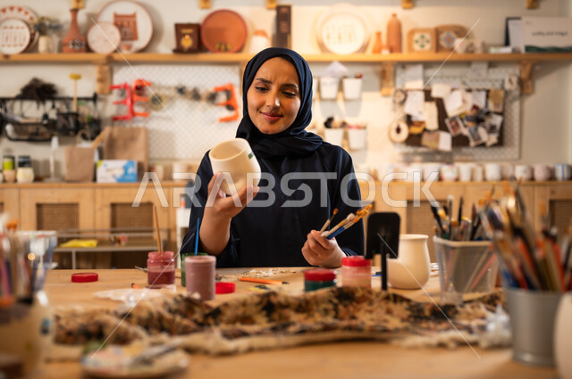  حرف ومهن شعبية تراثية قديمة, العمل في صناعة الخزفيات للاحتفال باليوم الوطني السعودي, امرأة عربية خليجية سعودية محجبة ترتدي العباءة السوداء تحمل بيدها معدات الرسم, إحياء ذكرى تأسيس الدولة السعودية 1727 م