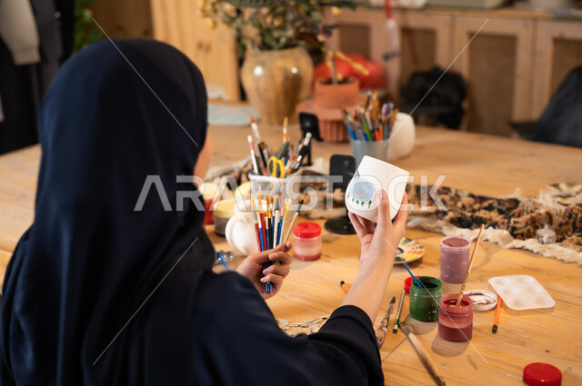 Working in the ceramics industry, the founding of the Saudi state in 1727 AD, the anniversary of Saudi National Day, September 23, the art of drawing and coloring on ceramics, a veiled Saudi Gulf Arab woman wearing a black abaya drawing on ceramics