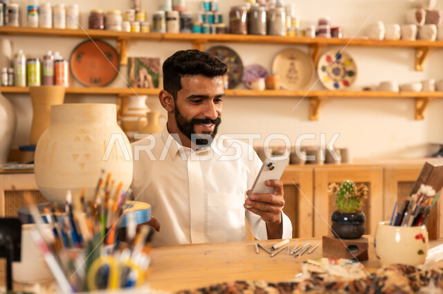 Sculptures, decorations and handmade artworks, ancient traditional folk crafts and professions, commemorating the founding of the Saudi state in 1727 AD, celebrating Saudi National Day, handmade plastic industries, a young Saudi Gulf Arab man wearing traditional clothing holding a mobile phone in his hand