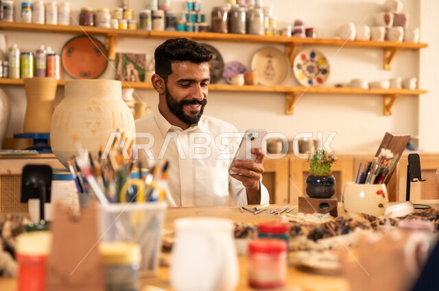 منحوتات وزخارف وأعمال فنية يدوية، حرف ومهن شعبية تراثية قديمة، إحياء ذكرى تأسيس الدولة السعودية 1727 م, الاحتفال باليوم الوطني السعودي, الصناعات التشكيلية اليدوية، شاب عربي خليجي سعودي يرتدي الثوب التقليدي يحمل بيده الجوال