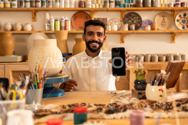 منحوتات وزخارف وأعمال فنية يدوية، حرف ومهن شعبية تراثية قديمة، إحياء ذكرى تأسيس الدولة السعودية 1727 م, الاحتفال باليوم الوطني السعودي, الصناعات التشكيلية اليدوية، شاب عربي خليجي سعودي يرتدي الثوب التقليدي يحمل بيده الجوال