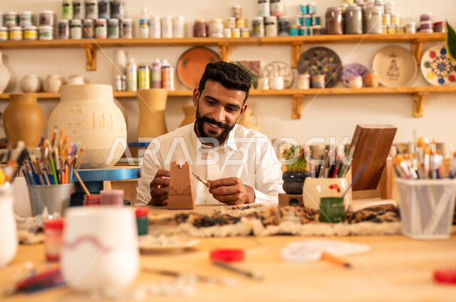 Sculptures, decorations and handmade artworks, ancient traditional folk crafts and professions, the founding of the Saudi state in 1727 AD, the anniversary of Saudi National Day September 23, handmade plastic industries, a young Saudi Gulf Arab man wearing traditional clothing drawing on pottery