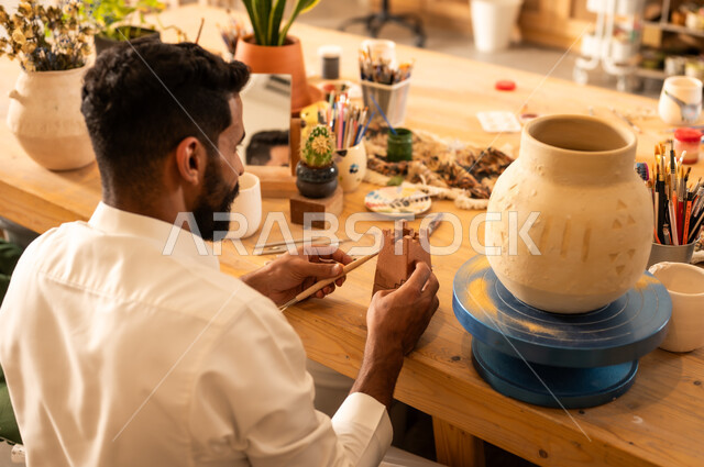 منحوتات وزخارف وأعمال فنية يدوية، حرف ومهن شعبية تراثية قديمة، تأسيس الدولة السعودية 1727 م, ذكرى اليوم الوطني السعودي 23 سبتمبر, الصناعات التشكيلية اليدوية، شاب عربي خليجي سعودي يرتدي الثوب التقليدي يقوم بالرسم على الفخار