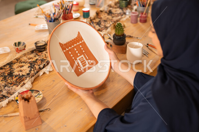 العمل في صناعة الخزفيات, فن الرسم والتلوين على الخزف، امرأة عربية خليجية سعودية محجبة ترتدي العباءة السوداء تحمل بيدها رسوم لبيت طيني باللون البني على طبق من السيراميك, تأسيس الدولة السعودية 1727 م, ذكرى اليوم الوطني السعودي23 سبتمبر