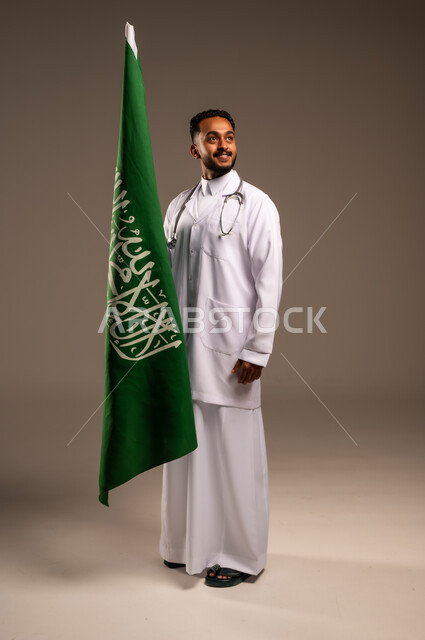 مهن ووظائف سعودية طبية، مفهوم العمل في القطاع الطبي، بورتريه لطبيب عربي خليجي سعودي يرتدي الثوب التقليدي والزي الطبي والسماعات يحمل بيده علم المملكة، إحياء ذكرى العيد الوطني السعودي 23سبتمبر، الإحتفال بيوم العلم 11 مارس، الطول الكامل للجسم، خلفية رمادية 
