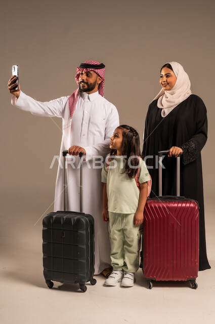 Taking family selfies, getting ready for a fun summer vacation, portrait of a Saudi Arabian Gulf man holding a mobile phone and taking a photo with his family next to suitcases, gestures of happiness and joy for going on a tourist trip, gray background