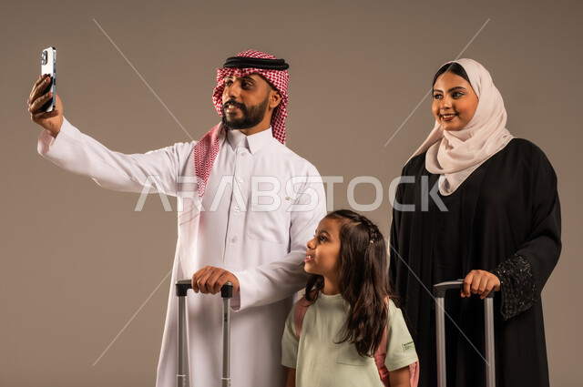 Taking family selfies, getting ready for a fun summer vacation, portrait of a Saudi Arabian Gulf man holding a mobile phone and taking a photo with his family next to suitcases, gestures of happiness and joy for going on a tourist trip, gray background