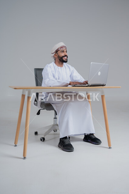 استخدام الأجهزة الحديثة والتقنيات المتطورة، مفهوم ادارة الاعمال عن بعد، مهن ووظائف مكتبيه عمانية، بورتريه لشاب عربي خليجي عماني يرتدي الدشداشة والعمامة يجلس خلف طاولة خشبية وامامه حاسوب محمول، خلفية بيضاء