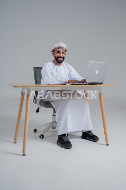 استخدام الأجهزة الحديثة والتقنيات المتطورة، مفهوم ادارة الاعمال عن بعد، مهن ووظائف مكتبيه عمانية، بورتريه لشاب عربي خليجي عماني يرتدي الدشداشة والعمامة يجلس خلف طاولة خشبية وامامه حاسوب محمول، خلفية بيضاء