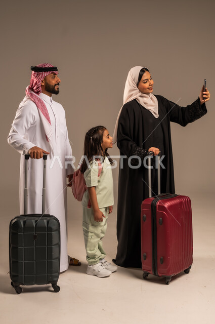  الاستعداد لقضاء إجازة صيفية ممتعة، التقاط صور السيلفي التذكارية العائلية، بورتريه لامراة عربية خليجية سعودية تحمل الهاتف المحمول وتأخذ صورة برفقة عائلتها بجانب حقائب السفر، ايماءات السعادة والفرح للذهاب برحلة سياحية، خلفية رمادية