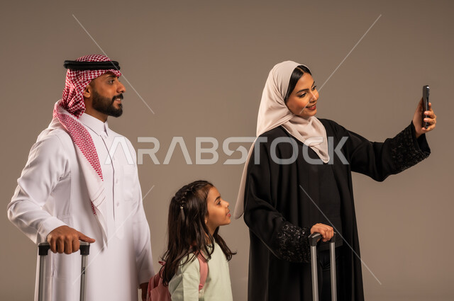  الاستعداد لقضاء إجازة صيفية ممتعة، التقاط صور السيلفي التذكارية العائلية، بورتريه لامراة عربية خليجية سعودية تحمل الهاتف المحمول وتأخذ صورة برفقة عائلتها بجانب حقائب السفر، ايماءات السعادة والفرح للذهاب برحلة سياحية، خلفية رمادية
