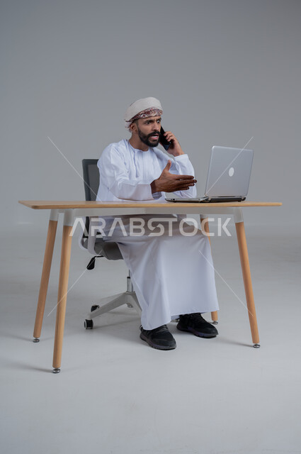ايماءات التعصيب والانفعال، استخدام الأجهزة الحديثة والتقنيات المتطورة، مفهوم ادارة الاعمال عن بعد، مهن ووظائف مكتبيه عمانية، بورتريه لشاب عربي خليجي عماني يرتدي الدشداشة والعمامة يجلس خلف طاولة خشبية وامامه حاسوب محمول يقوم بإجراء مكالمة هاتفية، خلفية بيضاء