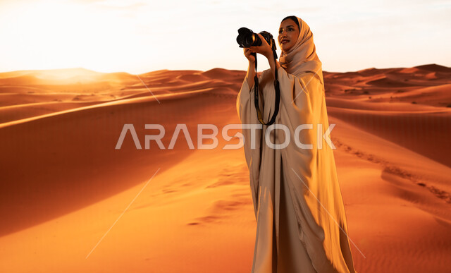 لقطات فنية فوتوغرافية، ممارسة الهواية المفضلة بشغف، مصورة محترفة تقوم بالتقاط الصور التذكارية باستخدام التقنيات الحديثة، الرمال الذهبية الناعمة, شابة عربية خليجية سعودية ترتدي عباءة ملونة تقف في المناطق الصحراوية تحمل بيديها كاميرا عالية الدقة