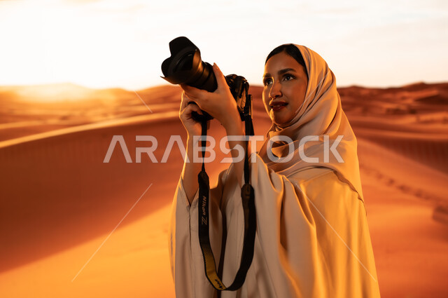 لقطات فنية فوتوغرافية، ممارسة الهواية المفضلة بشغف، مصورة محترفة تقوم بالتقاط الصور التذكارية باستخدام التقنيات الحديثة، الرمال الذهبية الناعمة, شابة عربية خليجية سعودية ترتدي عباءة ملونة تقف في المناطق الصحراوية تحمل بيديها كاميرا عالية الدقة