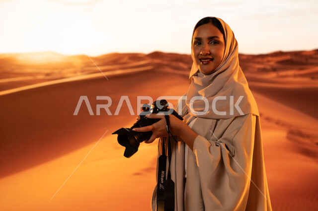 Artistic photography shots, practicing a favorite hobby with passion, a professional photographer taking souvenir photos using modern techniques, soft golden sand, a young Saudi Arabian Gulf woman wearing a colorful abaya standing in the desert areas holding a high-resolution camera in her hands